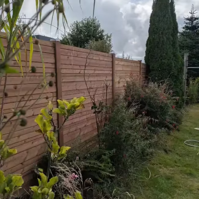 Google Rezension: Hoher Zaun aus Thermoholz als Sichtschutz, im Vordergrund Gartenpflanzen und im Hintergrund hohe Nadelbäume.